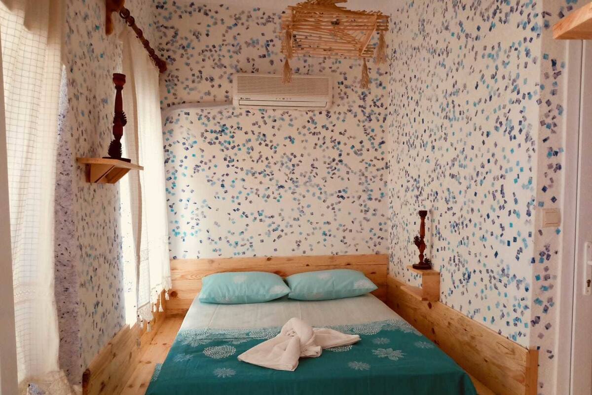 A bedroom with a wooden bed, teal bedding, a small bedside table, a wall-mounted air conditioner, and a window with a grid pattern.