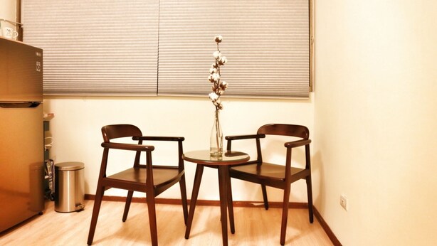 A small dining area with two wooden chairs and a round table, a trash can, and a window with blinds. - Good-Days Lovely Suite BRT Calligraphy Greenway (West Dist.)