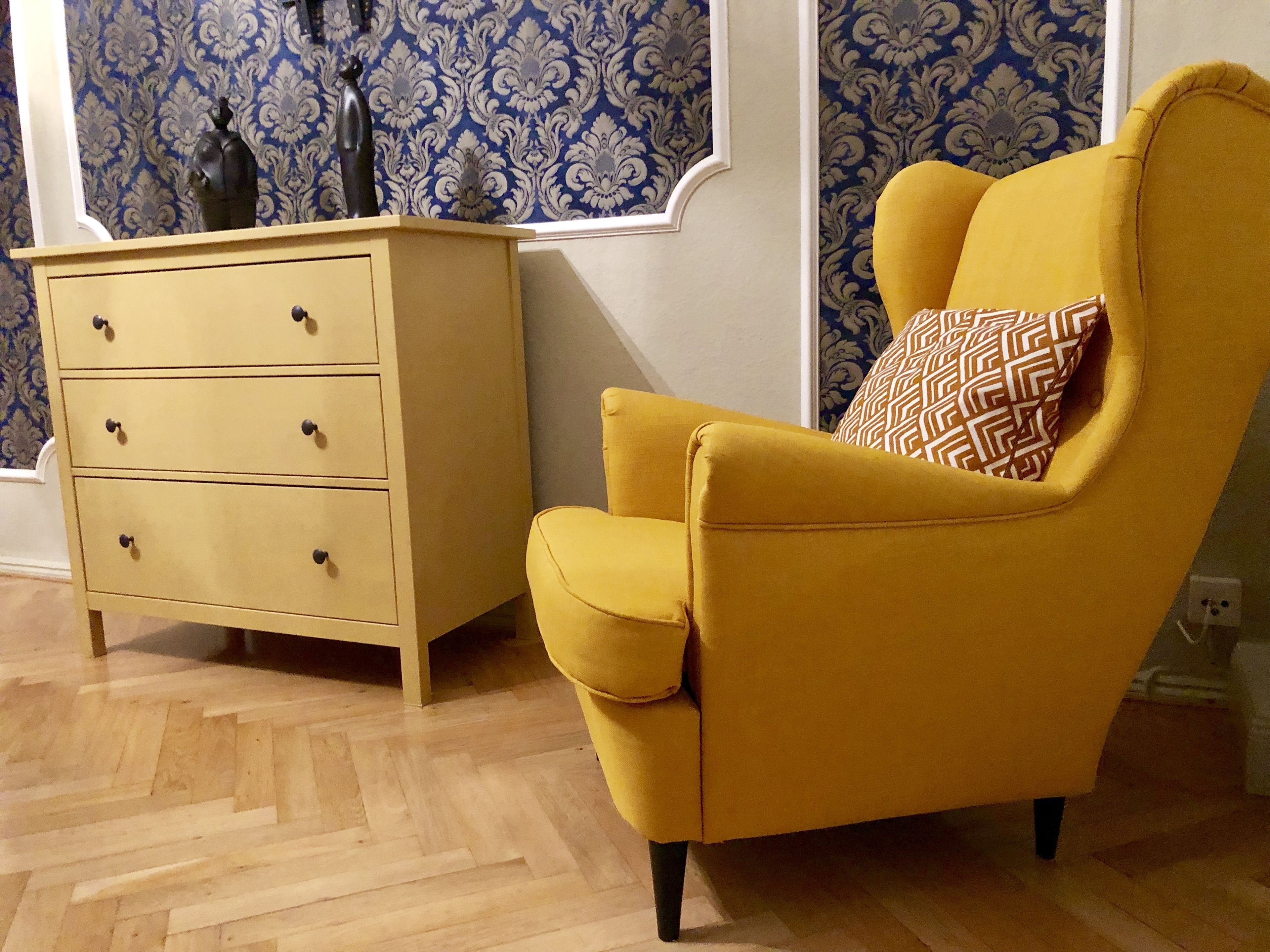 A yellow armchair with a patterned pillow, a wooden dresser with three drawers, and blue patterned wallpaper.