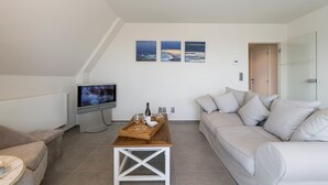 Una sala de estar moderna con un televisor de pantalla plana, una mesa de centro de madera con una bandeja, un sofá beige y un sofá blanco. Hay tres cuadros en la pared.