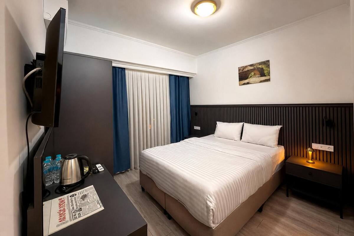 Ett hotellrum med en säng, ett skrivbord med en kaffebryggare, en TV, ett nattduksbord med en lampa och ett fönster med gardiner.