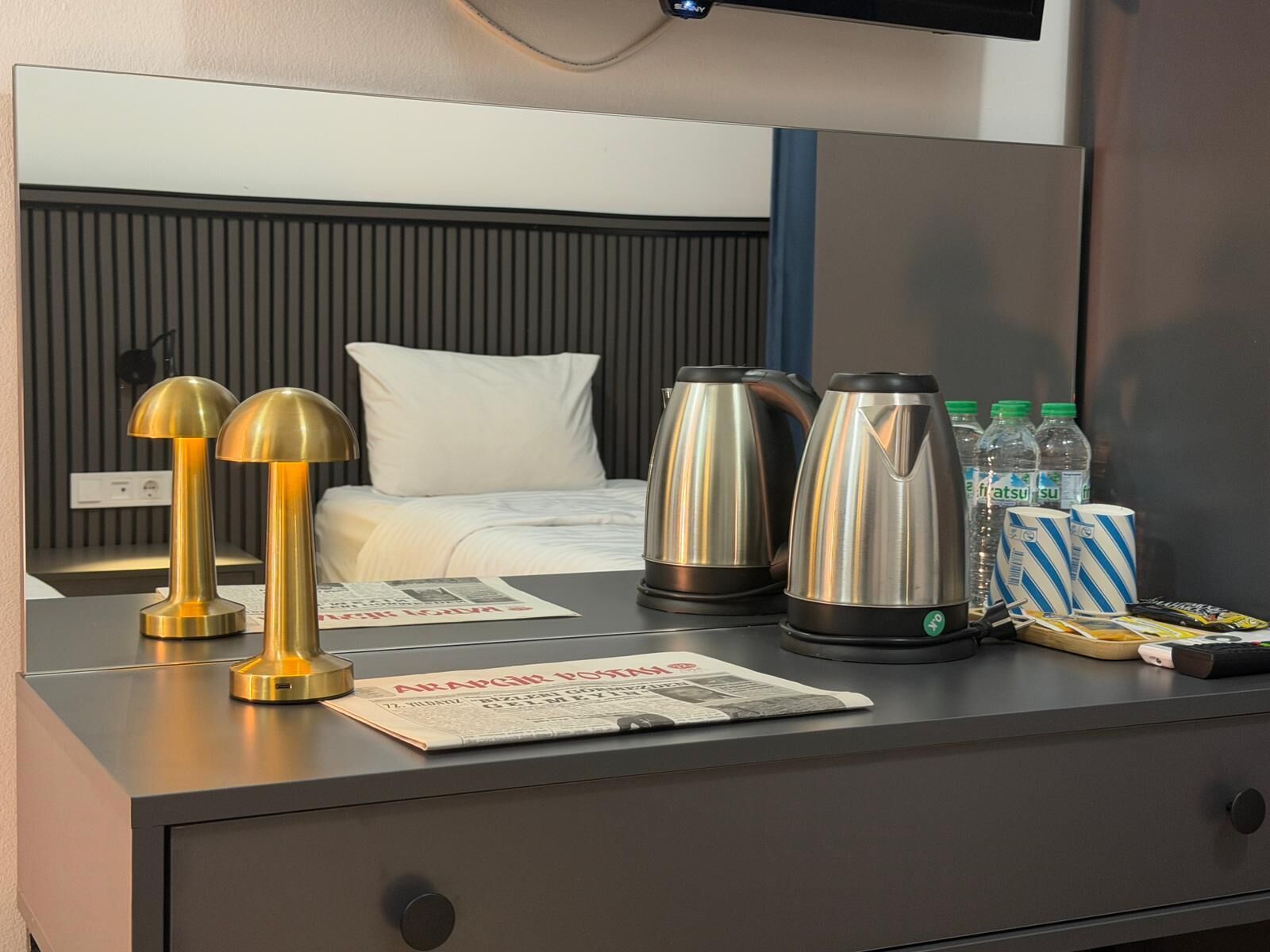 Una habitación de hotel con cama, lámparas de noche, periódico, tetera y agua embotellada.