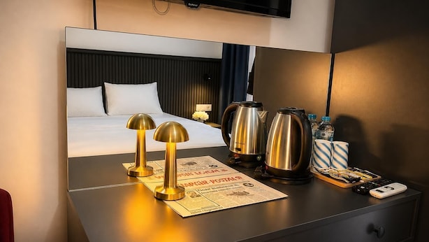 Une chambre d’hôtel avec un lit, une table de chevet sur laquelle repose un journal, deux bouilloires électriques, une télécommande et un téléphone intelligent.