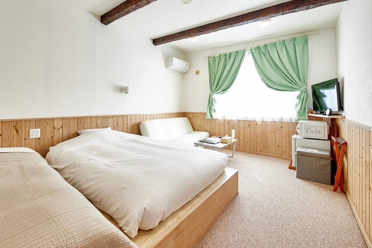 A bedroom with a wooden headboard, a bed with white bedding, a bedside table, a TV, and a window with green curtains.