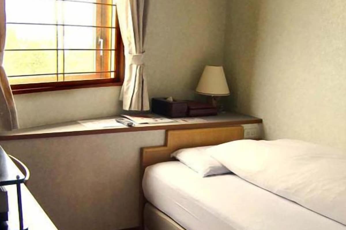 Une chambre d’hôtel avec un lit, une fenêtre avec des rideaux, une table de chevet avec une lampe et une petite table avec un livre.