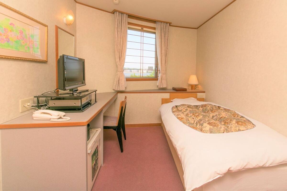 Una habitación de hotel con cama, escritorio, computadora, teléfono, ventana con cortinas y un cuadro colgado en la pared.