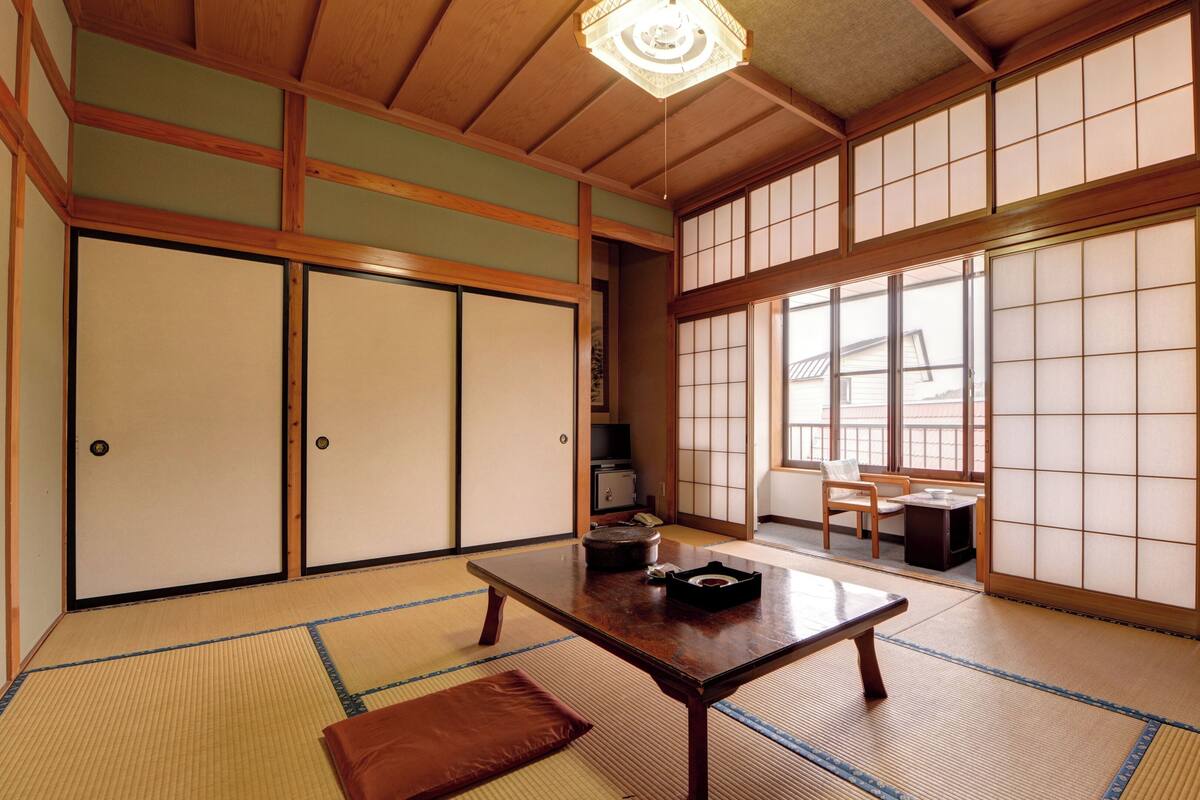 Une pièce de style japonais traditionnel, avec un sol recouvert de tatamis, des portes coulissantes, une table en bois et une vue dégagée sur l’extérieur grâce à de grandes fenêtres.