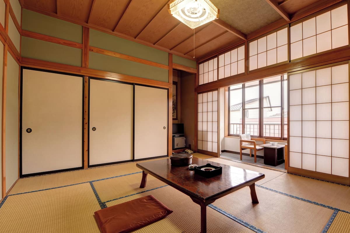 A traditional Japanese room with tatami flooring, sliding doors, a wooden table, and a view through large windows.