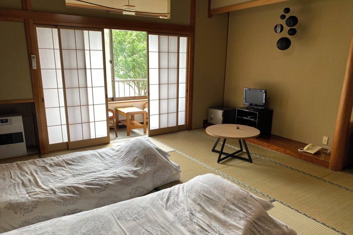 Une pièce de style japonais traditionnel, avec un sol recouvert de tatamis, deux futons posés sur des tatamis, une petite table ronde, un téléviseur et une porte coulissante donnant sur un balcon.