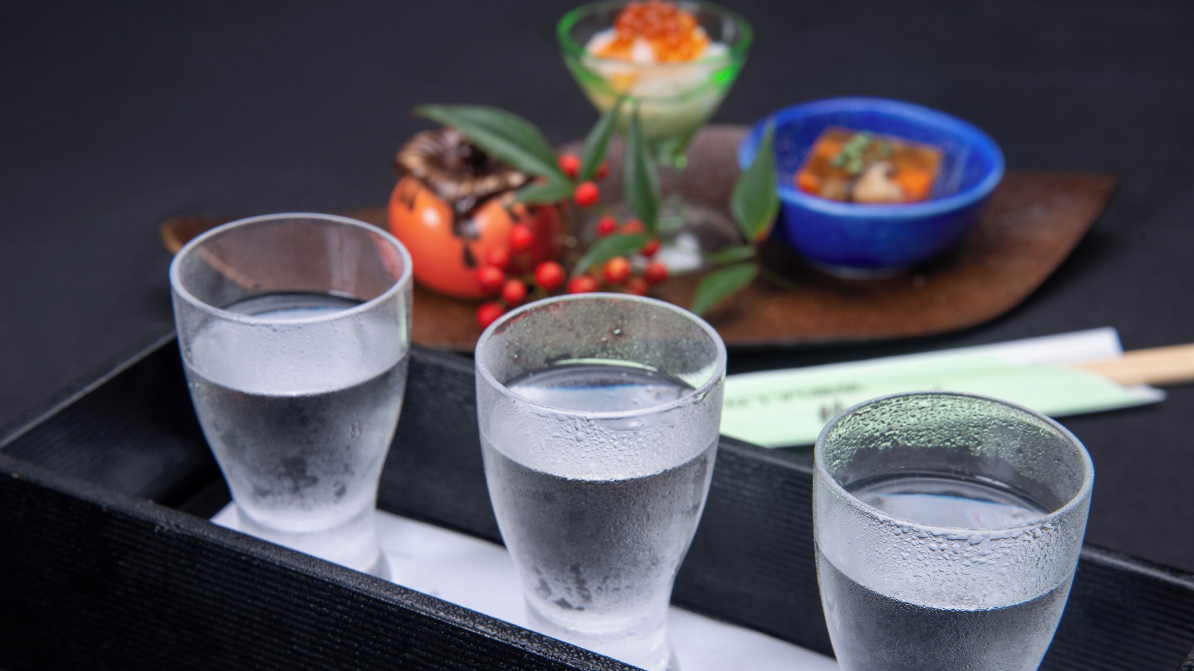 Three glasses of water on a tray with a wooden board, a bowl of food, and a small dish with red berries.