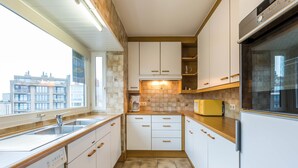 A modern kitchen with white cabinets, a built-in oven, and a window overlooking a cityscape. - Panisa 401 (Koksijde)