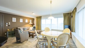 A modern living room with a round dining table, wicker chairs, a sofa, and a television. - Panisa 401 (Koksijde)