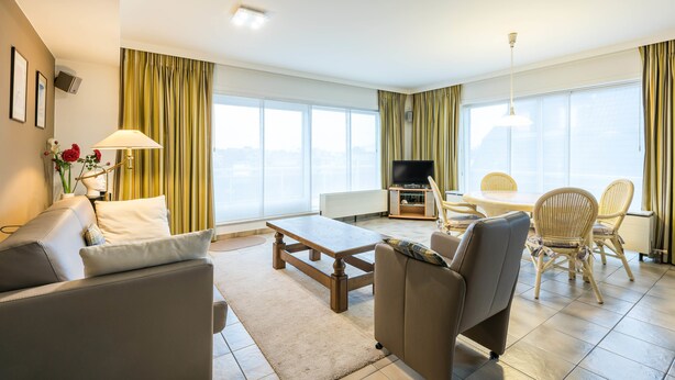A modern living room with a sofa, armchair, dining table, and chairs. There is a television and a window with curtains. - Panisa 401 (Koksijde)