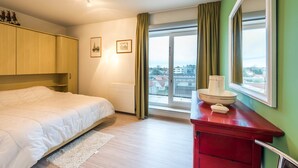 A hotel room with a bed, a red dresser, a window with a view, and a small table with a bowl and a cup. - Panisa 401 (Koksijde)