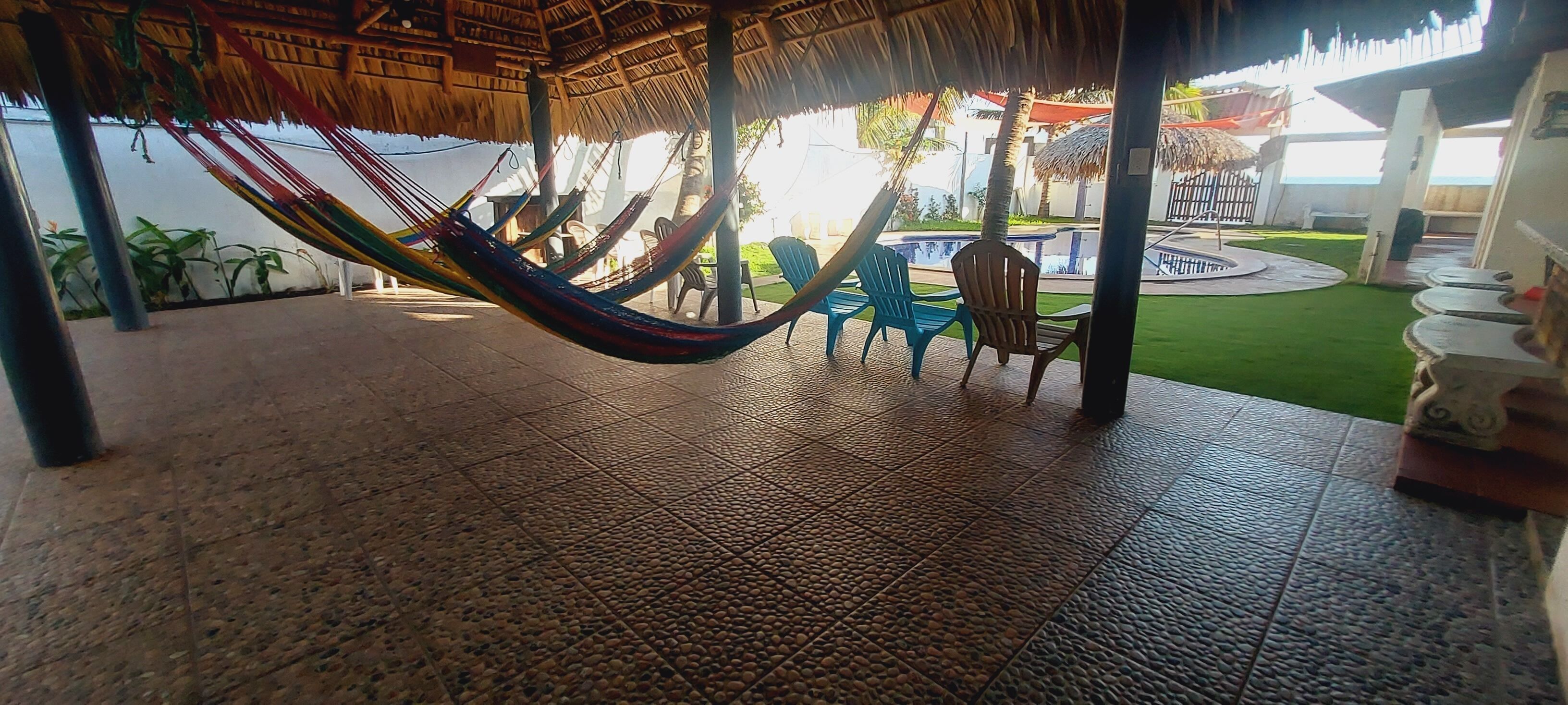 A hammock under a thatched roof, outdoor chairs, and a pool area.