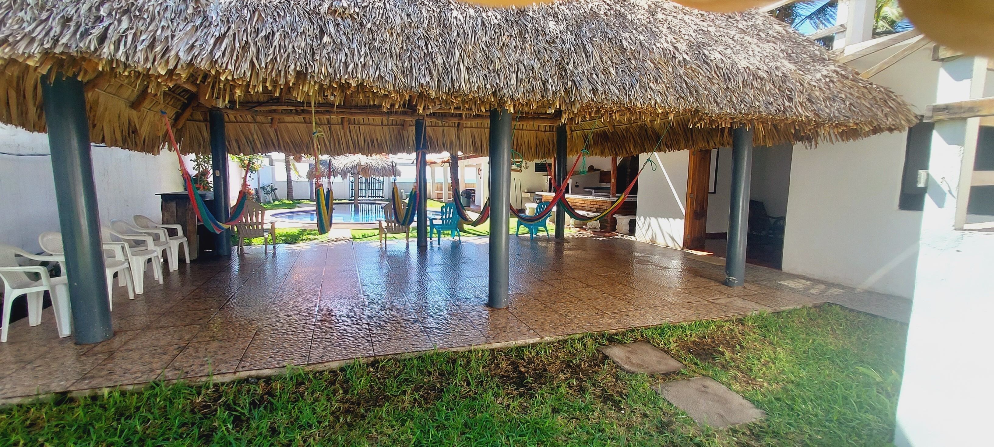 A covered outdoor seating area with thatched roofs, plastic chairs, and a pool in the background.