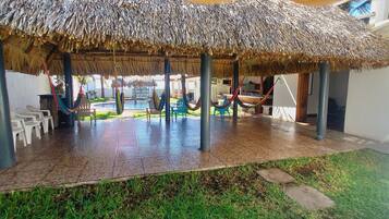 A covered outdoor seating area with thatched roofs, plastic chairs, and a pool in the background.