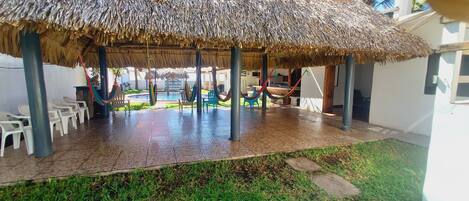 A covered outdoor seating area with thatched roofs, plastic chairs, and a pool in the background.