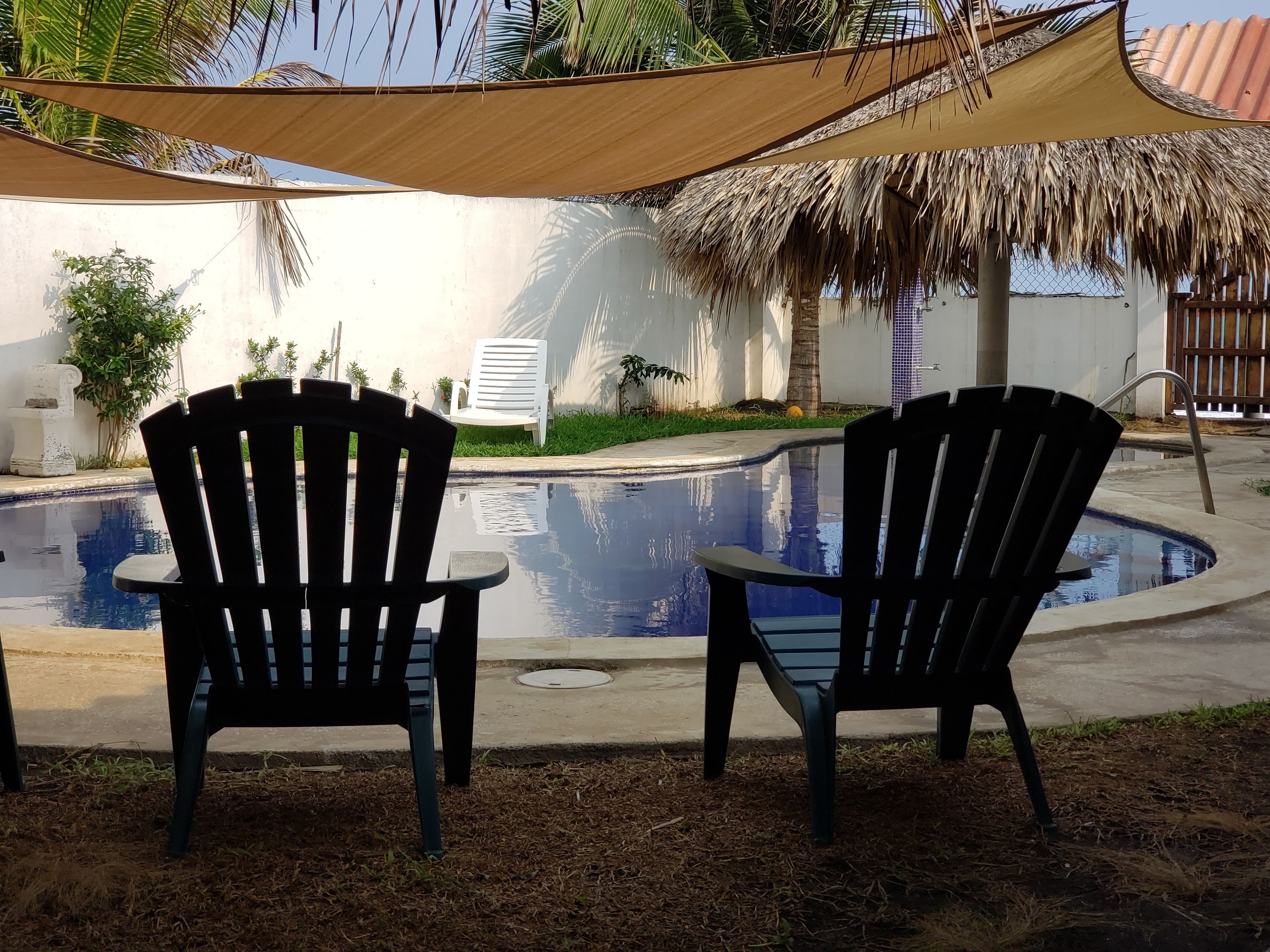 Two Adirondack chairs facing a swimming pool with a thatched roof and palm trees in the background.