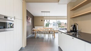 A modern kitchen with white cabinets, a black countertop, and a dining area with wooden chairs and a table. - Koetje Boe (Koksijde)