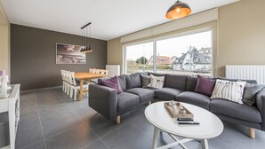 A modern living room with a grey sectional sofa, a round coffee table, and a dining area with a wooden table and chairs. - Koetje Boe (Koksijde)