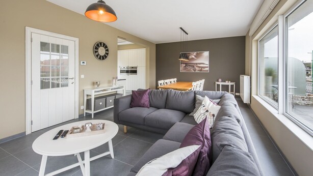 A modern living room with a grey sofa, a white coffee table, and a dining area with a wooden table and chairs. - Koetje Boe (Koksijde)