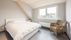 A bedroom with a sloped ceiling, a large bed, a wicker chair, and a window with a view of buildings. - Koetje Boe (Koksijde)