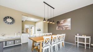 A modern kitchen with a wooden table and white chairs, a clock, pendant lights, and a painting on the wall. - Koetje Boe (Koksijde)
