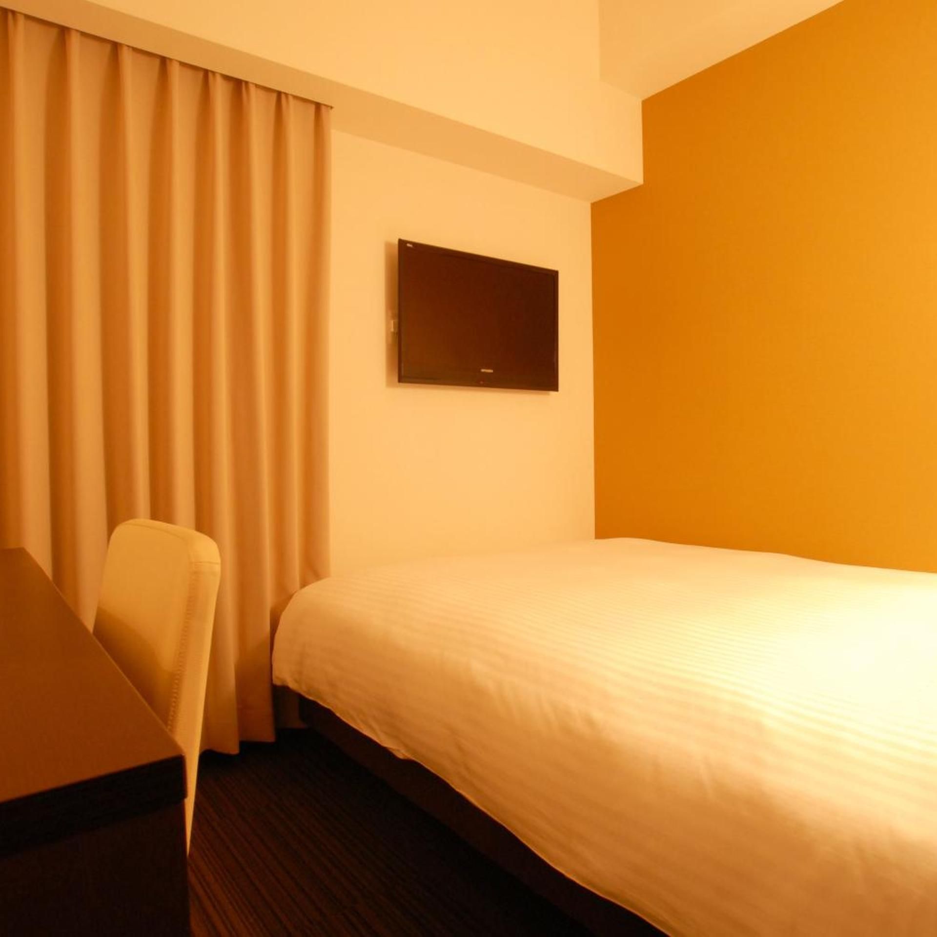 A hotel room with a bed, a chair, a flat-screen TV mounted on the wall, and a window with curtains.
