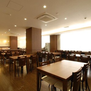 A spacious dining area with multiple wooden tables and chairs, a kitchen area in the background, and a large window providing natural light. - Ab Hotel Komaki (Komaki)