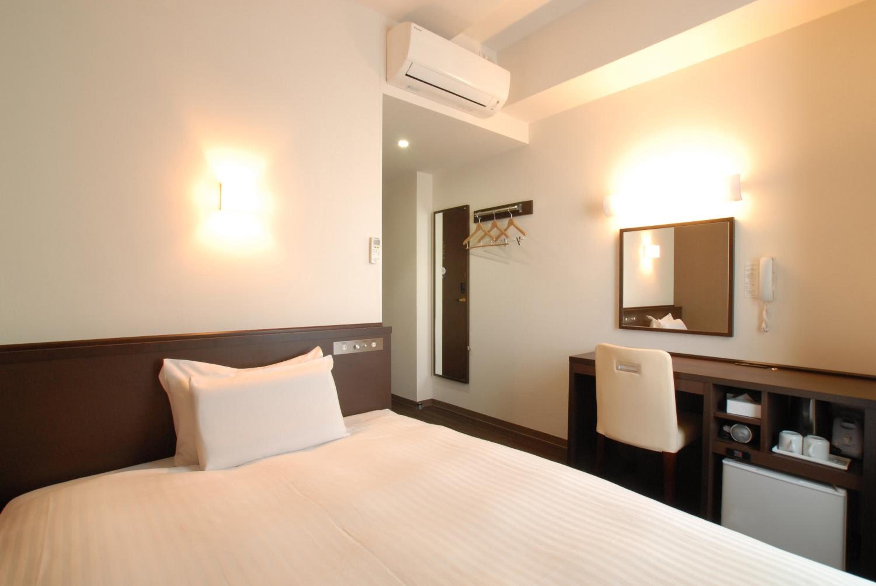 A hotel room with a bed, a desk, a mirror, and an air conditioning unit.