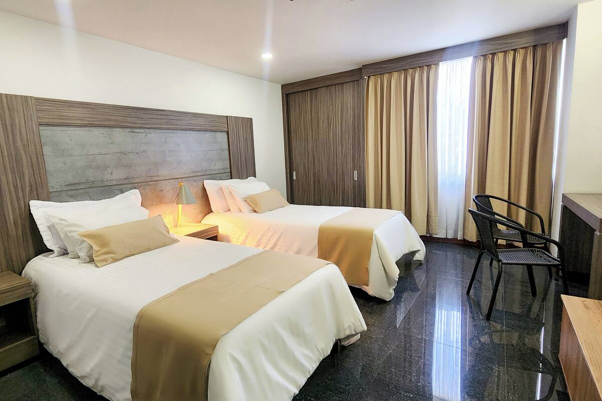 A hotel room with two beds, a desk, a chair, and a large window with curtains.