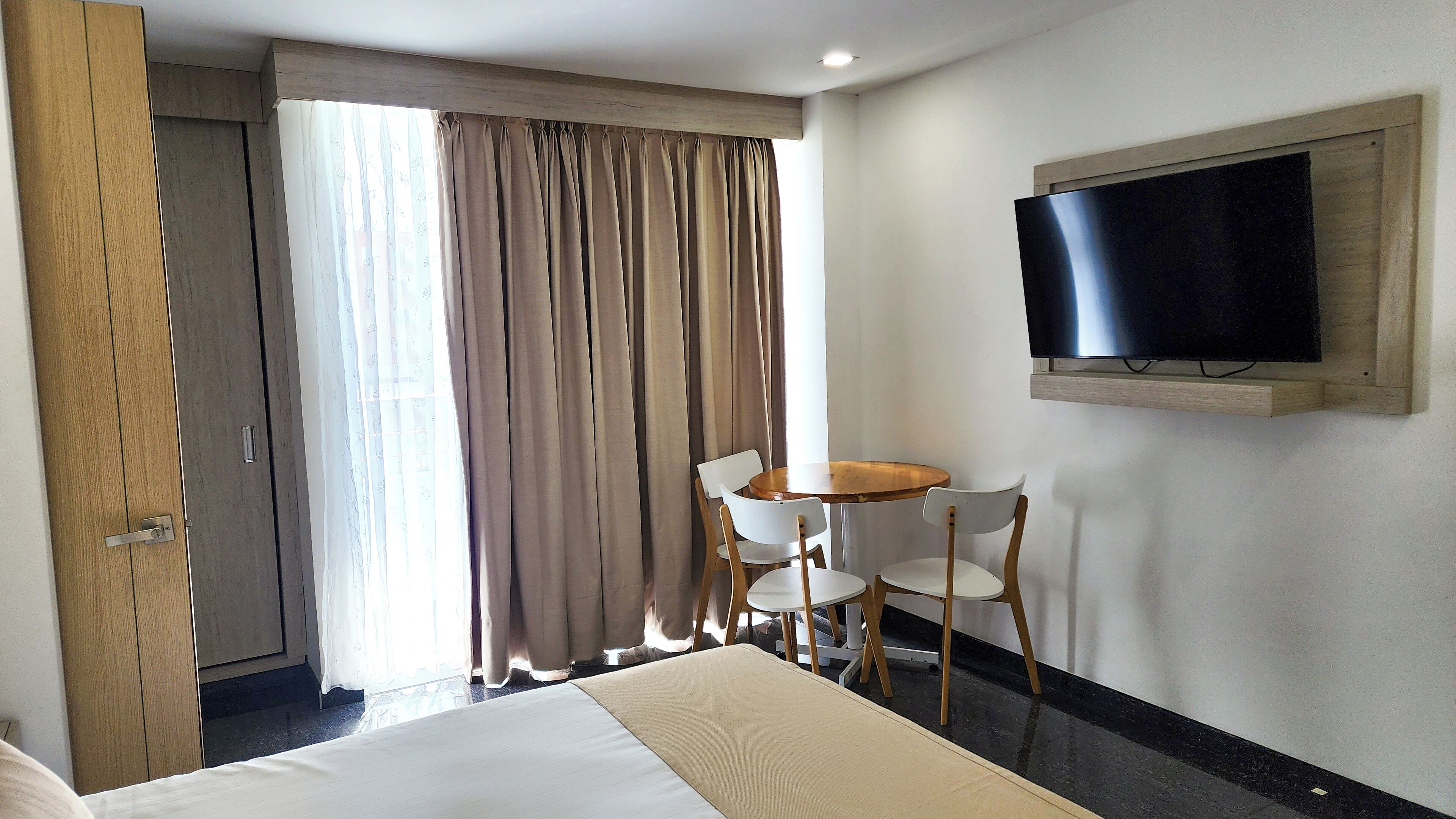 Habitación de hotel con cama, televisor montado en la pared, una mesita con dos sillas y una ventana con cortinas.
