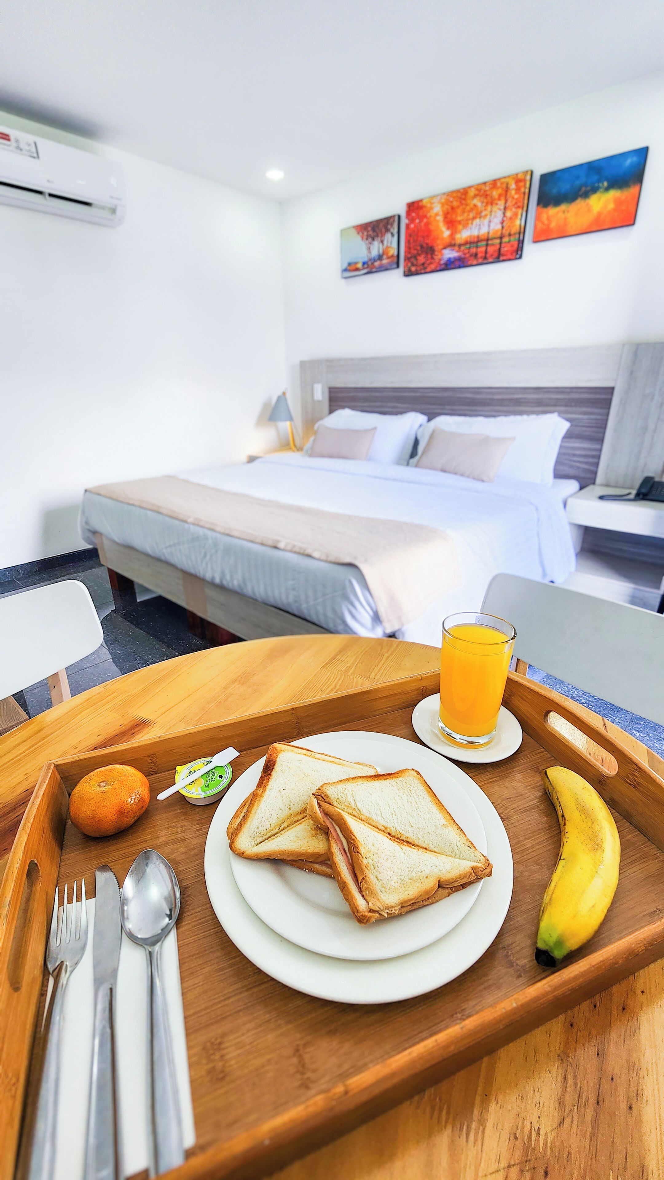 A hotel room with a bed, a bedside table, a tray with breakfast items, and artwork on the wall.