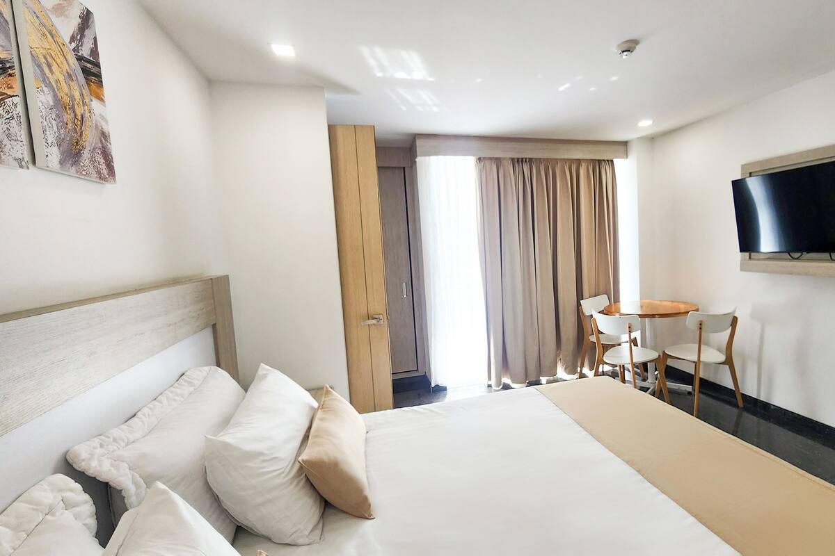A hotel room with a bed, a TV mounted on the wall, a small table with chairs, and a door leading to another room.