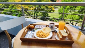 Bandeja de café da manhã com ovos mexidos, torradas, suco de laranja e uma caneca em uma varanda com vista para um jardim.