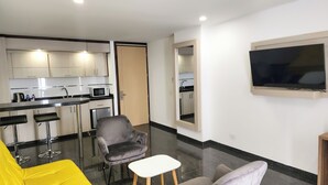 Um espaço de estar compacto com cozinha americana, uma poltrona cinzenta, uma mesa pequena branca e um sofá amarelo.