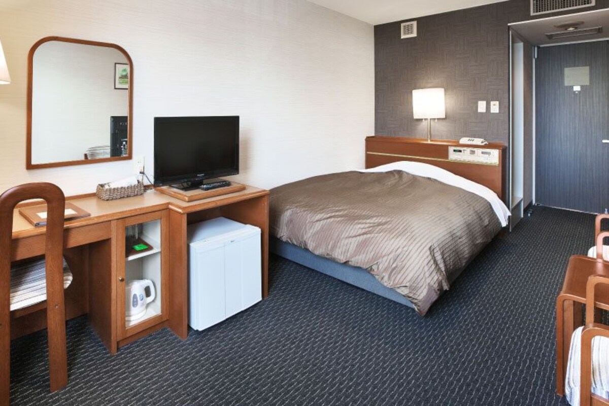 A hotel room with a bed, a desk with a computer, a chair, a small refrigerator, a mirror, and a door.