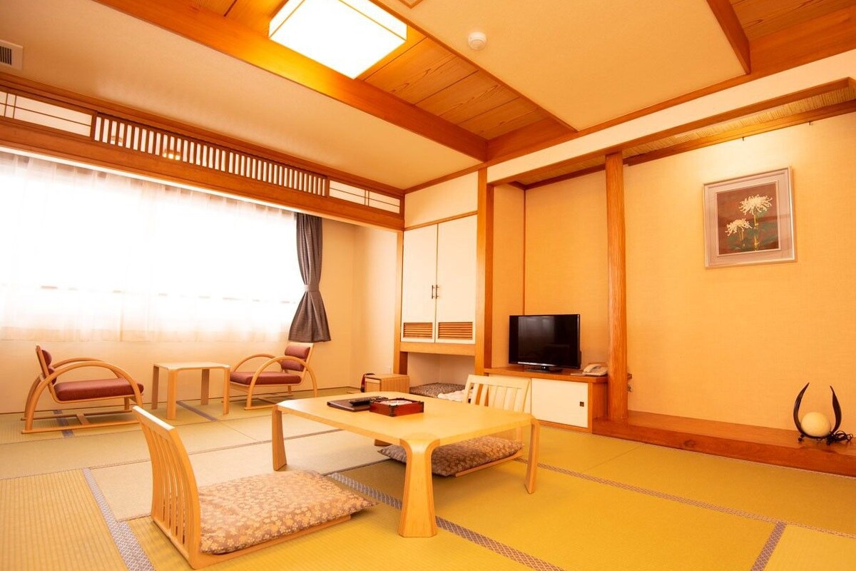 A traditional Japanese-style room with tatami flooring, a low table, cushioned seating, a television, and a cabinet.