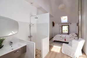 Casa de banho moderna com chuveiro, pia e espelho. No quarto vizinho, há uma cama e uma janela.