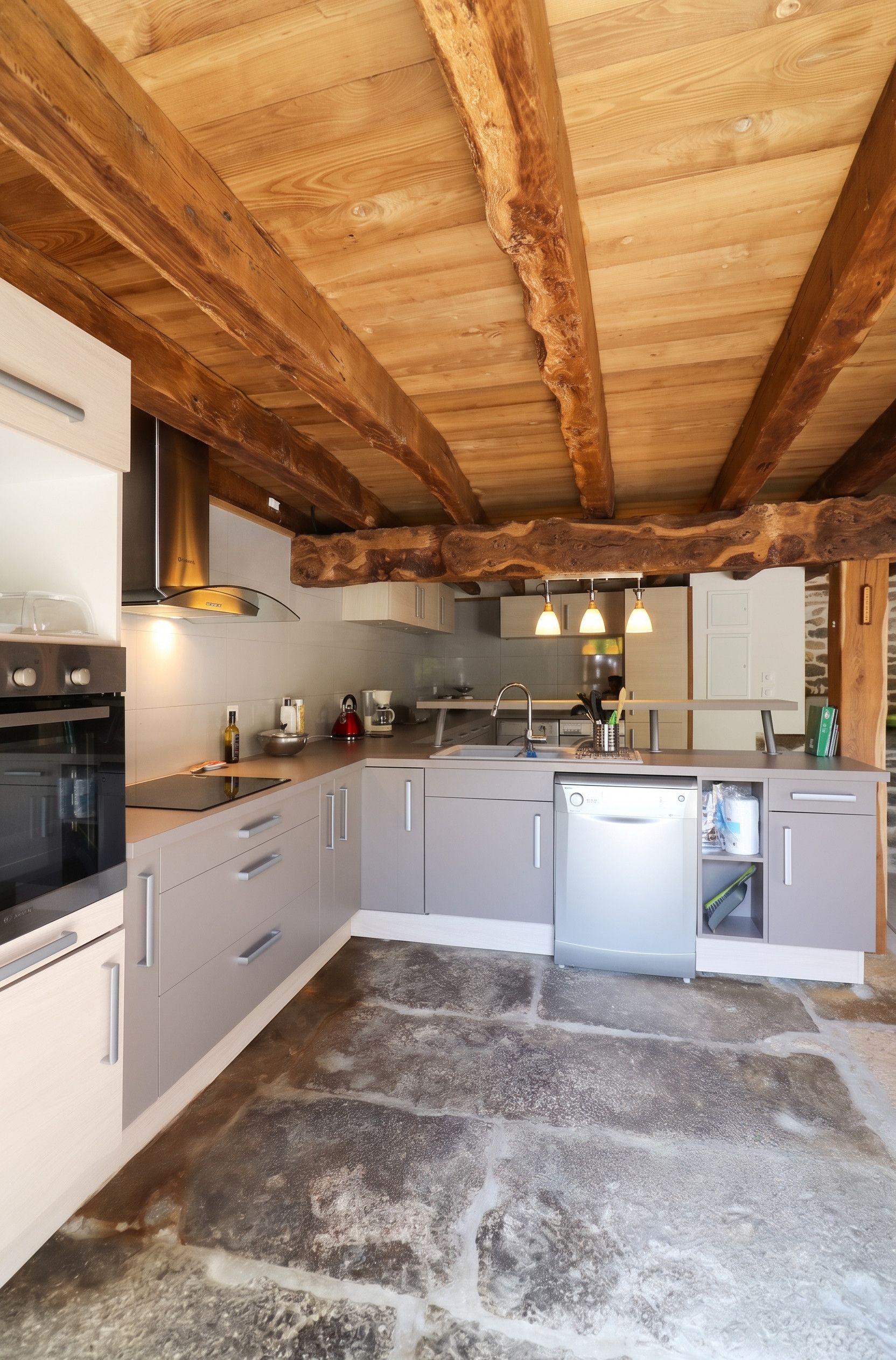 A modern kitchen with a stone floor, wooden beams, and stainless steel appliances.