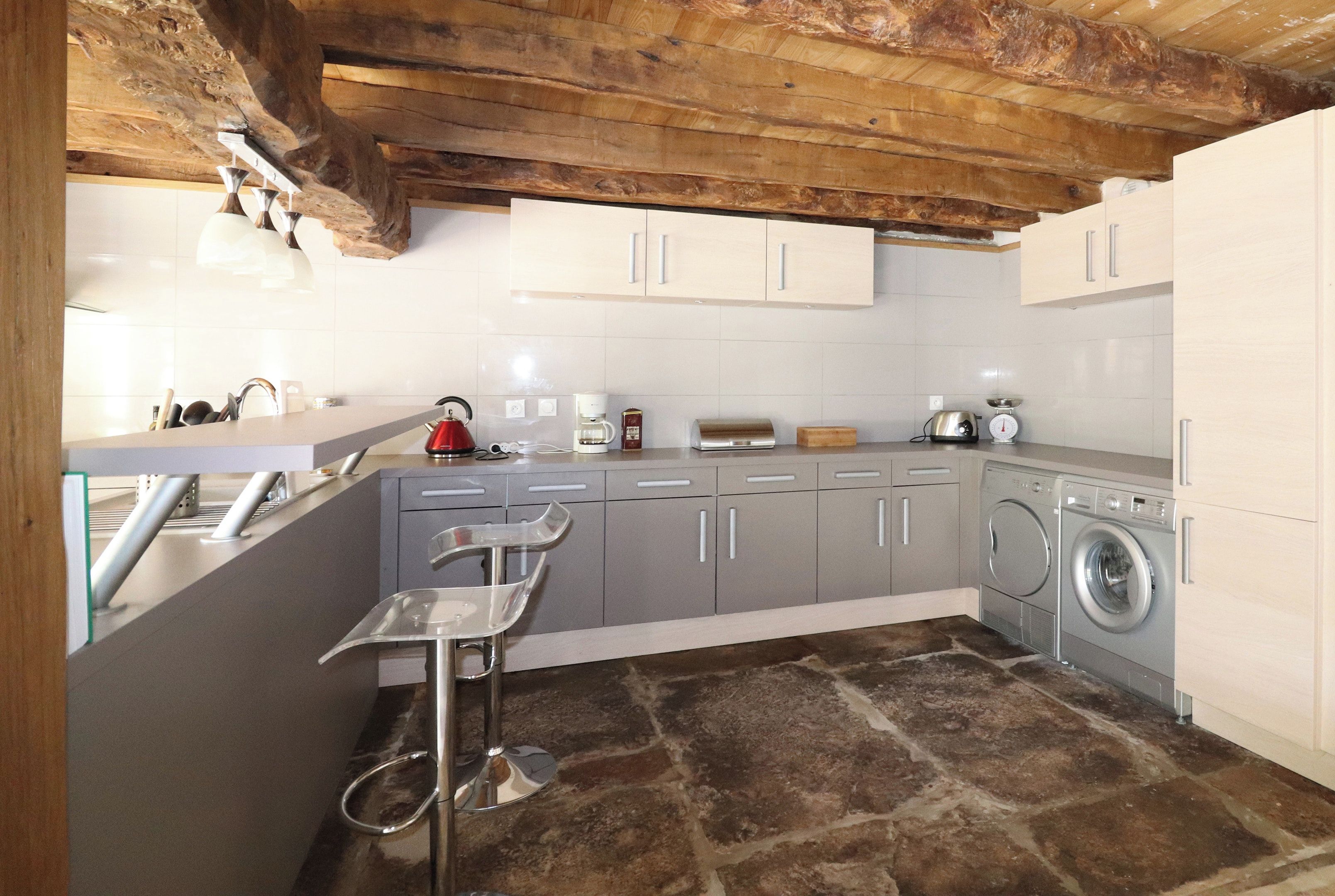 A kitchen with stone flooring, grey cabinets, a washing machine, and a bar stool.