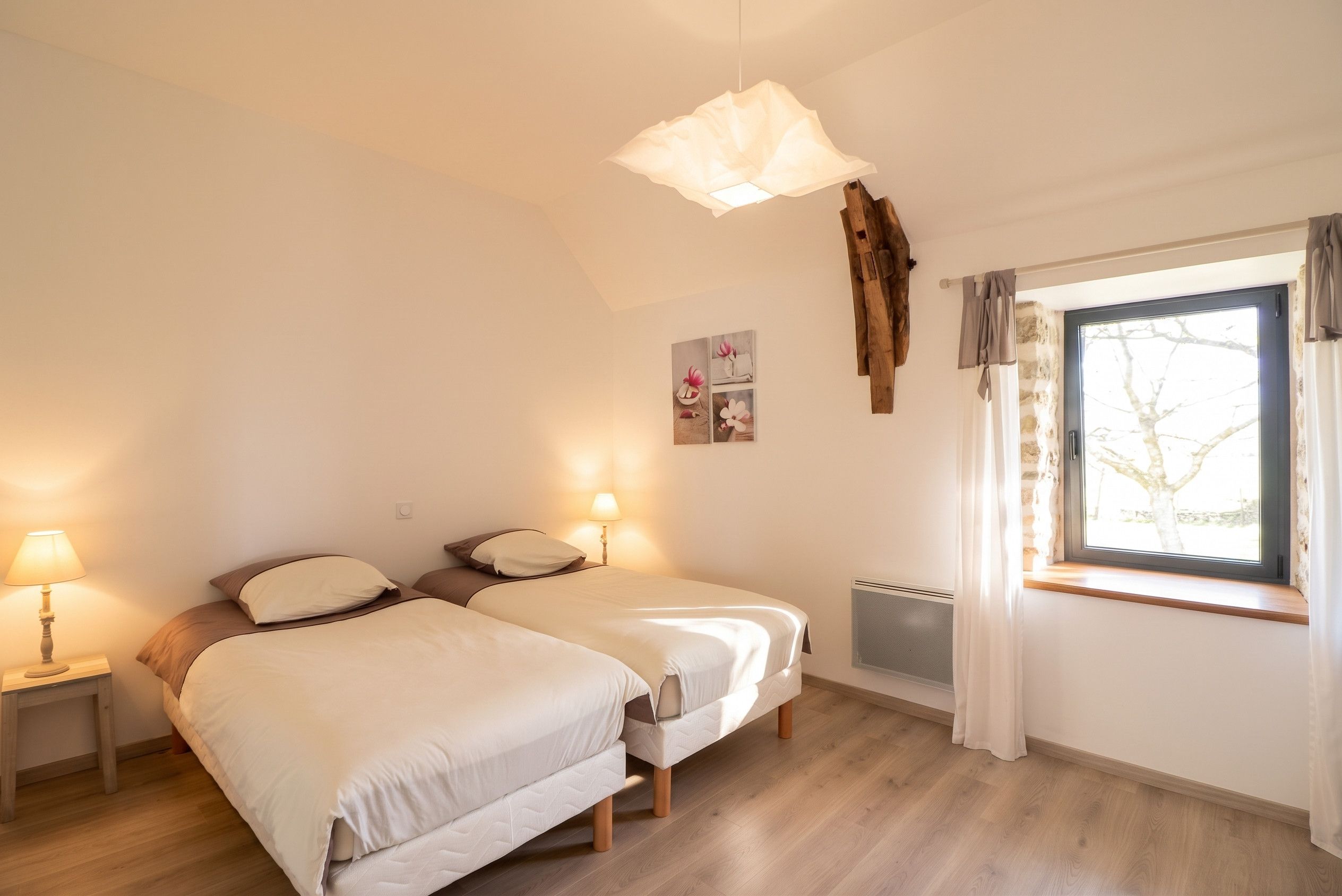 Ein Schlafzimmer mit zwei Betten, einem fensterbelaubten Fenster, einer Pendelleuchte und einer Heizkörper.