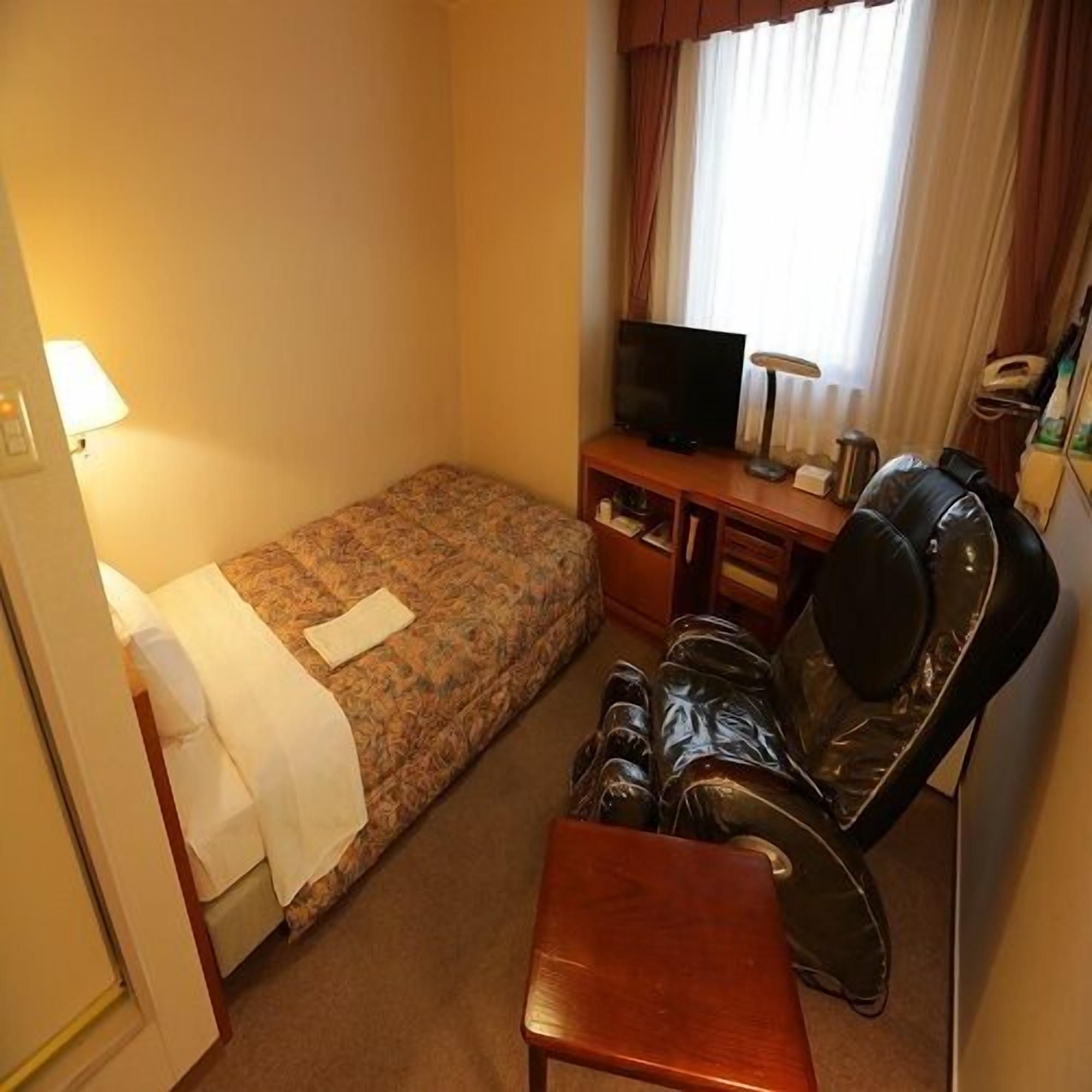 Een kleine hotelkamer met een eenpersoonsbed, een bureau met een televisie, een stoel en een lamp.