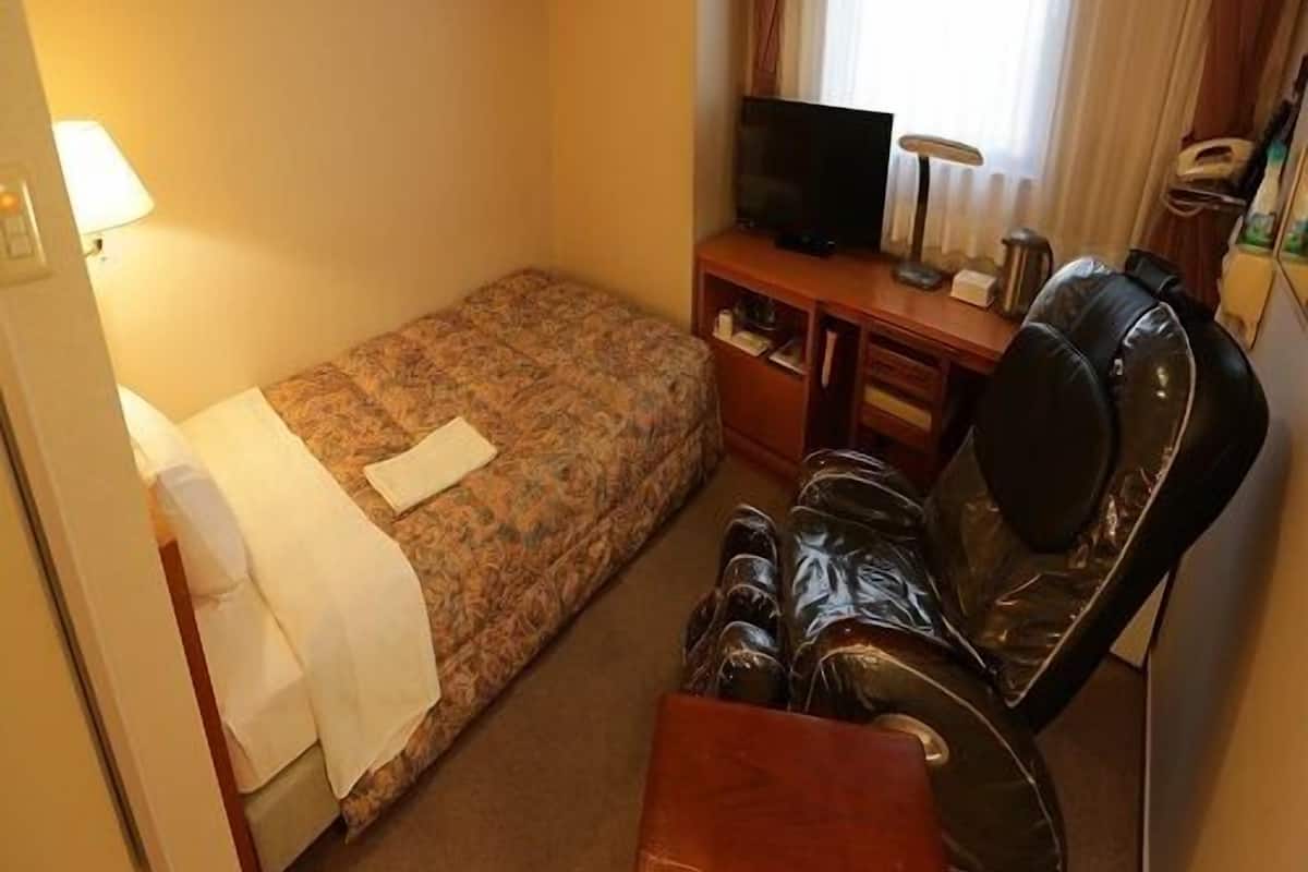 A small hotel room with a single bed, a desk with a TV, a chair, and a lamp.