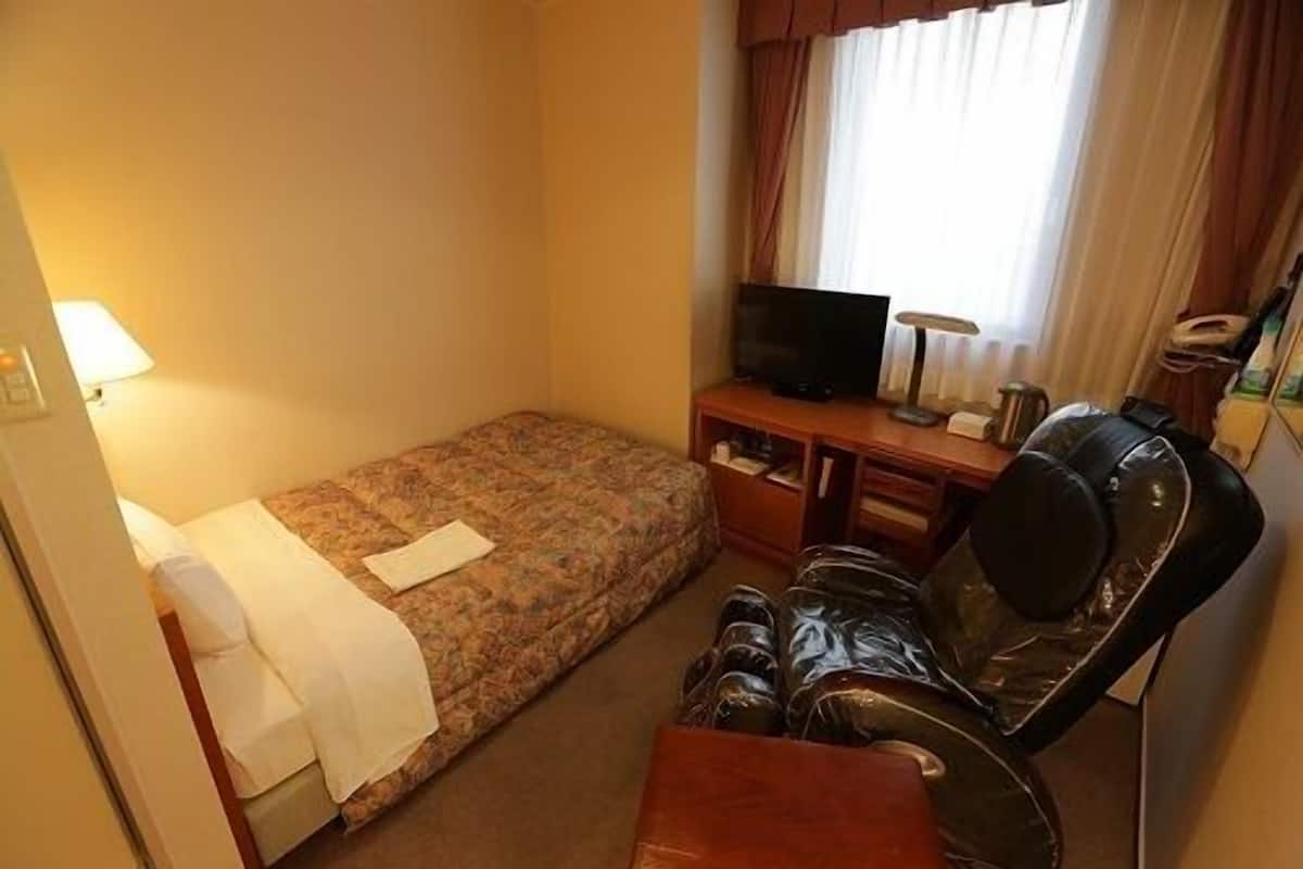 A compact hotel room with a single bed, a desk with a television, and a massage chair.