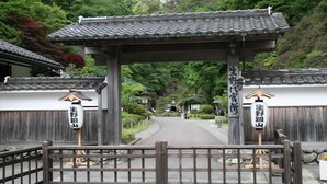 Ein traditionelles japanisches Tor mit hölzernen Säulen und einem Ziegeldach, das zu einem Pfad führt, der von Holzzäunen und Grünpflanzen gesäumt wird.