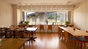 Ein Konferenzraum mit Holztischen und -stühlen, großen Fenstern mit Blick auf eine Brücke und Grünflächen sowie Blumentöpfen auf der Fensterbank.