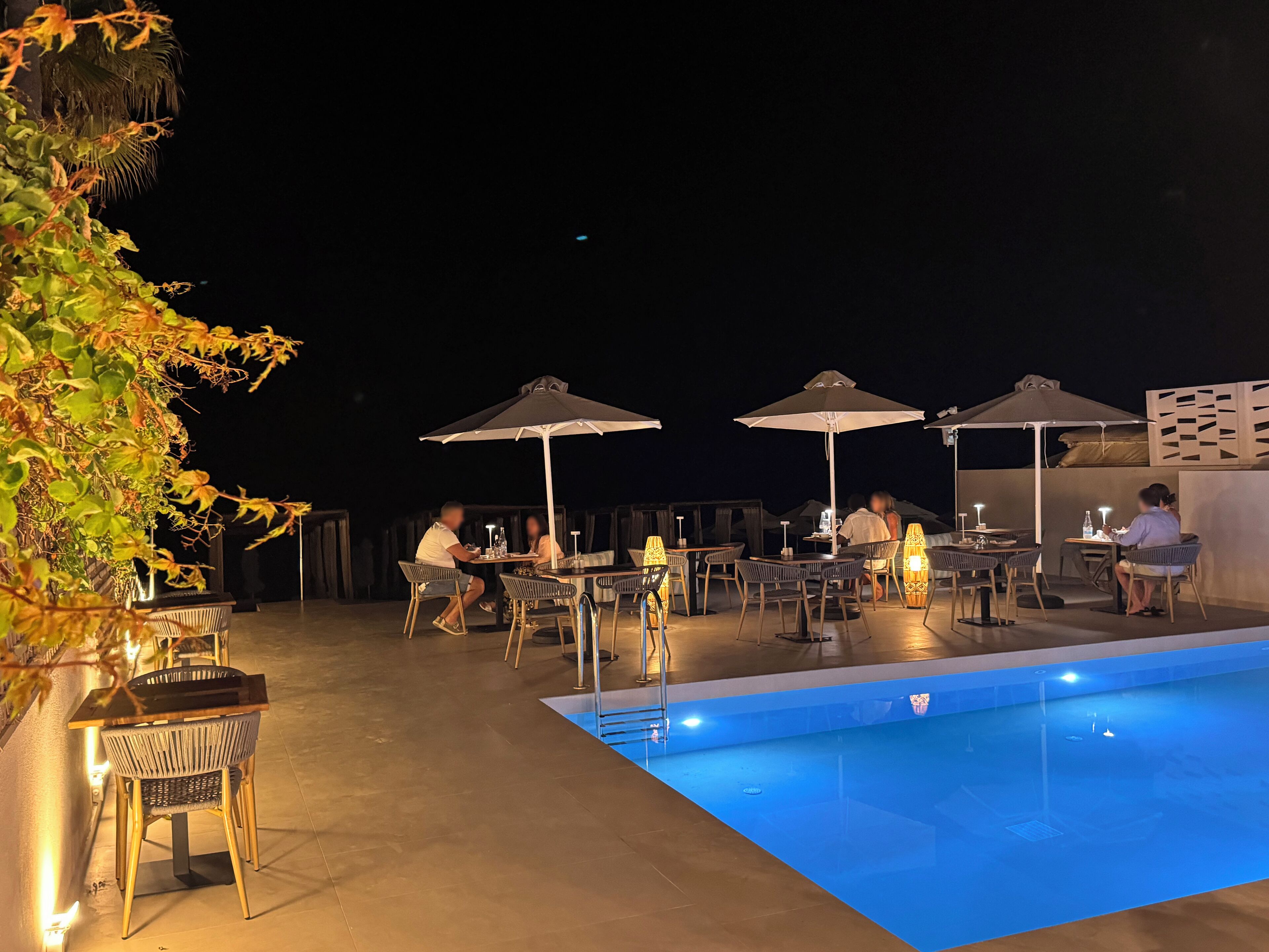 An outdoor pool area with tables and chairs under umbrellas, surrounded by greenery.