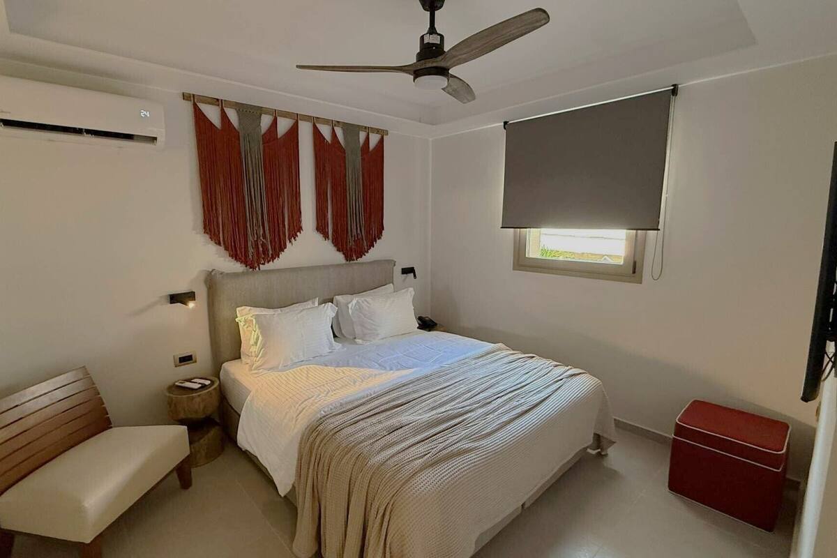 Une chambre avec un lit, un banc, un ventilateur de plafond, un appareil de climatisation, une fenêtre avec un store et un meuble rouge.